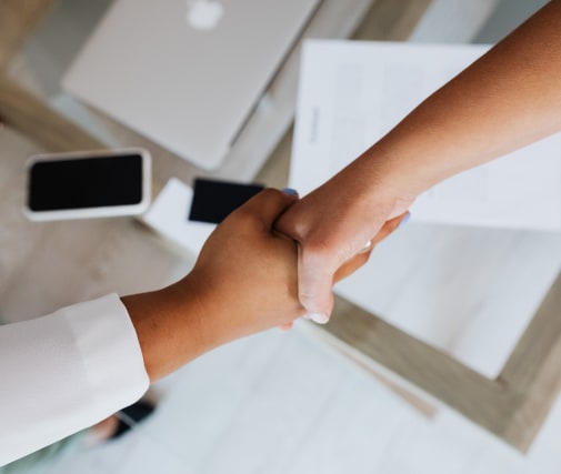 Two people shaking hands over a desk with documents, a laptop, and a smartphone visible.