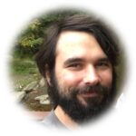 A bearded man with dark hair smiling outdoors, with greenery and rocks in the blurred background.