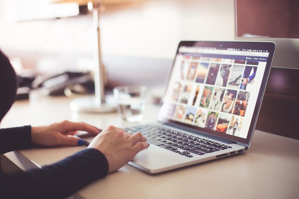 Person browsing photos on a laptop at a desk with a glass of water, lamp, and office supplies in the background.
