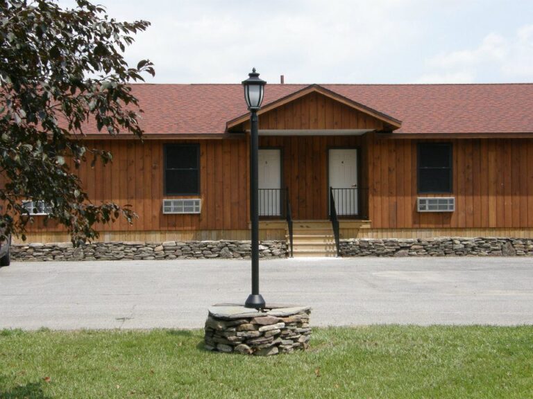 Wooden building with two white doors and a lamp post on a stone base in front, set on a grassy lawn.