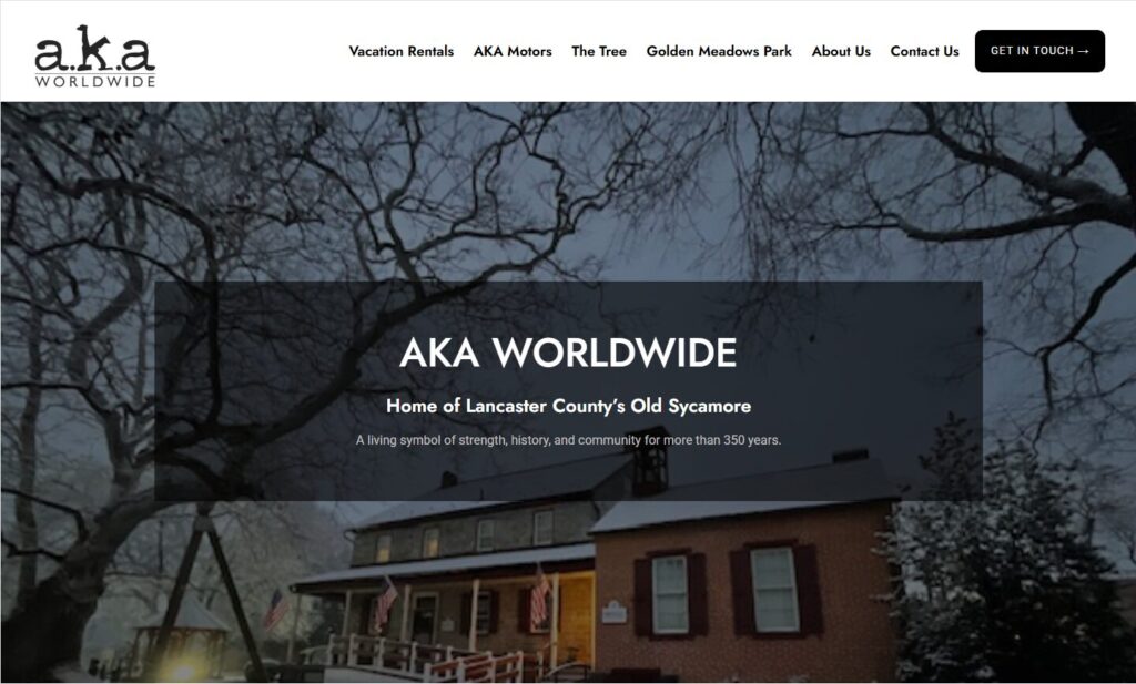 A large sycamore tree and historic brick house with AKA Worldwide website branding and navigation menu overlaid.