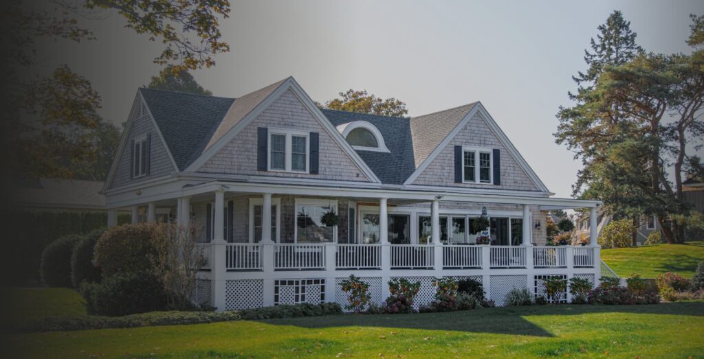 White two-story house with a wraparound porch, perfect for your website, nestled among a green lawn, trees, and shrubs.