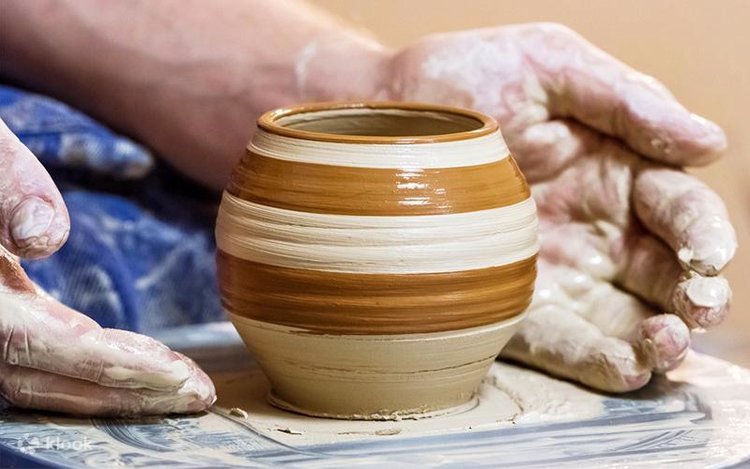 Hands shaping a striped clay pot on a pottery wheel for a website; the pot has brown and beige horizontal bands.