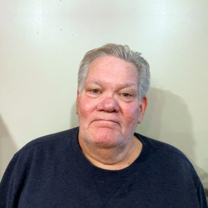 Older man with gray hair and a navy blue shirt for a website, standing against an off-white wall, looking at the camera.