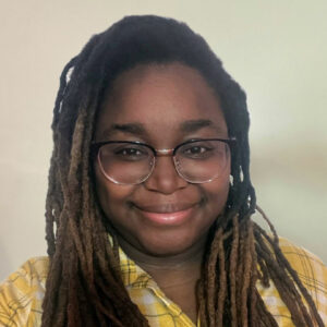 A person with long, dark dreadlocks and glasses smiles, perfect for a website, in a yellow plaid shirt against a light background.