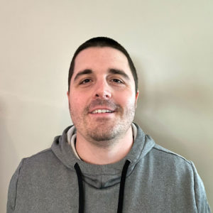 A man with short dark hair, slight stubble, and a gray hoodie smiles in front of a beige wall for a website photo.