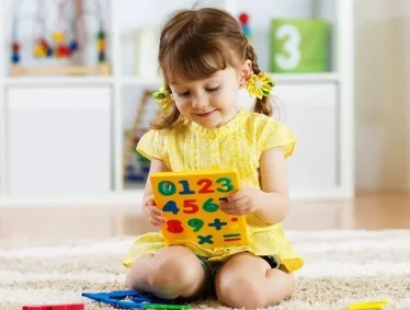 Young girl in yellow dress sits on floor, exploring a colorful math learning board with numbers for a website.