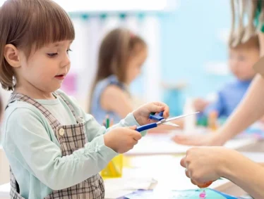 Young child holding scissors, cutting paper at a table; other children craft in the background for website use.