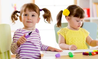 Two young girls sit at a table drawing with markers and modeling clay, perfect for a website featuring creative kids’ activities.