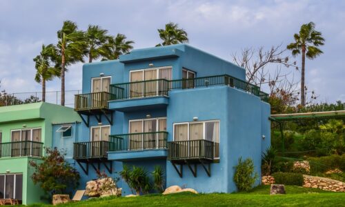 Blue two-story house with balconies, perfect for a website backdrop, surrounded by greenery and palm trees under a partly cloudy sky.