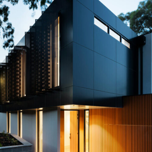 Modern two-story house with dark panels, vertical wood slats, large windows, and exterior lighting featured on our website.