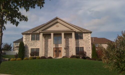 Two-story stone house with columns and large windows, perfect for a real estate website, sits under a partly cloudy sky.